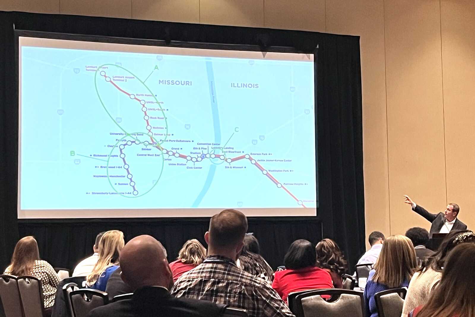 A speaker at a podium gestures at a large projection screen showing the MetroLink system map during a presentation to a group